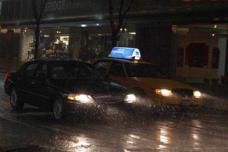 A night scene of a black town car and a yellow taxicab next to each other on a rain soaked street